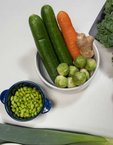 pexels photo 6811336 6811336 Top view of fresh vegetables including cucumbers, carrot, ginger, and edamame on a white surface.